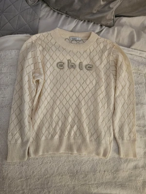 Jättegullig beige stickad tröja med pärldetaljer - Jättegullig beige stickad tröja från Fullamoda med diamantmönster och pärlbroderade bokstäver 'chic' framtill. Rund hals och långa ärmar. Aldrig använd och därför i mycket bra skick. 💓