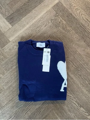 Marinblå stickad tröja från Ami - Säljer en marinblå stickad tröja från Ami med vit hjärtformad A-logga på bröstet. Tröjan har rund hals, ribbade muddar och långa ärmar. Perfekt för dig som gillar stilrena detaljer och vill ha något snyggt och bekvämt.