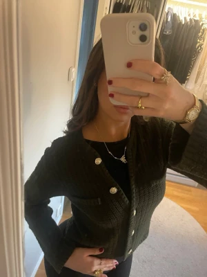 Mörkgrön stickad kofta med guld knappar - Snygg mörkgrön stickad kofta med guldiga knappar framtill och två små fickor på bröstet. Koftan har en croppad passform och långa ärmar, perfekt att styla med höga jeans eller kjol. Materialet är grovstickat och ger en mysig känsla.