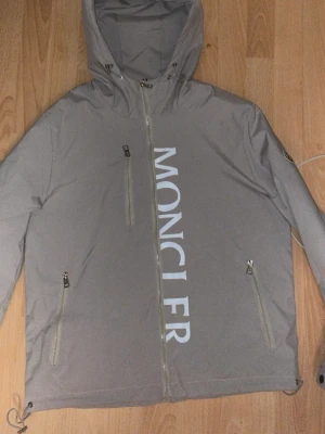 Grå Moncler vindjacka - Snygg grå vindjacka från Moncler med stor logga framtill och emblem på ärmen. Jackan har huva med snörning, flera dragkedjefickor och justerbar nederkant. Insidan är fodrad med silkesliknande material och har Moncler-märke. Perfekt för dig som gillar stilren design.