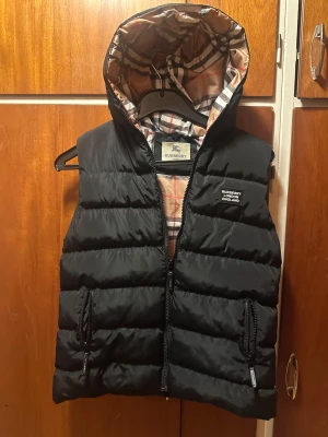 Svart dunväst från Burberry med huva - Svart dunväst från Burberry Yttermaterial i polyester och innerfoder i bomull. Perfekt för lager-på-lager och snygg stil. Storlek 13-14/small. Jag är runt 180cm och den är lite kort för mig. Hör av dig vid några frågor