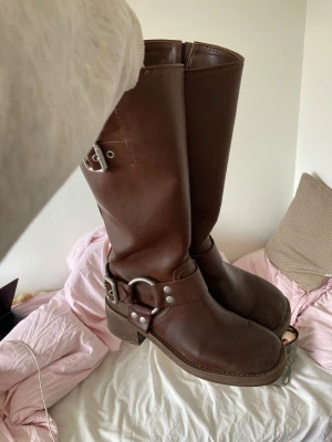 Steve madden Bruna biker boots med spännen - Säljer ett par snygga bruna biker boots i skinn med robust klack och rund tå. Bootsens höga skaft har coola metallspännen och ringdetaljer som ger en edgy vibe. Perfekta för dig som gillar en tuffare stil och vill sticka ut. Inköpspris:1700kr