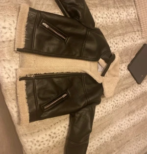 Svart teddyjacka från Bershka - Säljer en svart teddyjacka från Bershka med mjukt beige foder och detaljer som bälte vid midjan och dragkedjor på fickorna. Jackan har en klassisk krage och är tillverkad i syntetmaterial med skinnlook och teddyfoder. Perfekt för kalla dagar!