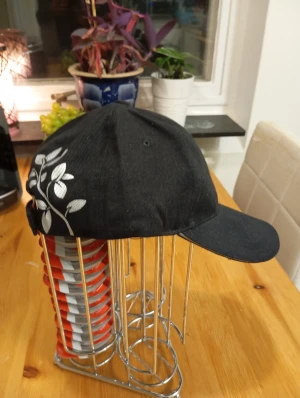 Svart keps med vit blommig detalj - Snygg svart keps med vit blommig print på sidan. Klassisk form med böjd skärm och justerbar rem bak. Perfekt för att lyfta din streetstyle eller casual look. Materialet känns som bomull och kepsen har en stilren vibe.