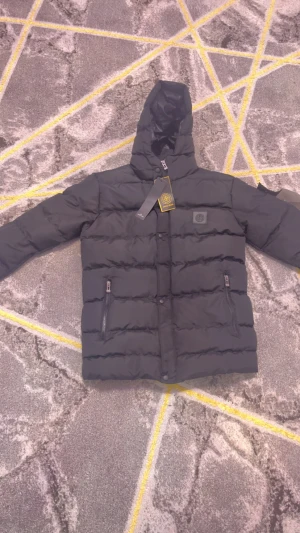 Svart pufferjacka från Stone Island - Svart pufferjacka från Stone Island med huva, två sidofickor med dragkedja och klassisk logotyp på bröstet. Jackan är tillverkad i polyester och har en clean, quiltad look med snygga detaljer. Perfekt för kalla dagar och har en modern, urban vibe.