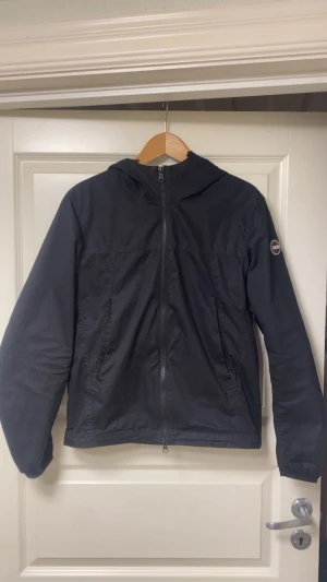 Svart vindjacka från Colmar, strl S - Svart vindjacka från Colmar Originals med huva och hel dragkedja framtill. Jackan har två sidofickor med dragkedja och Colmar-logga på vänster ärm. Tillverkad i lätt polyester, perfekt för blåsiga dagar. Enkel och stilren design som funkar till det mesta.