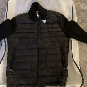 Svart pufferjacka från Moncler - Snygg svart pufferjacka från Moncler med dragkedja och långa ärmar. Jackan har en quiltad design på framsidan och ribbade muddar. Perfekt för kyligare dagar.