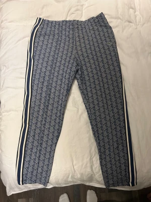 Blå mönstrade mjukisbyxor Lacoste XL - Snygga blå mjukisbyxor från Lacoste med geometriskt mönster och vita sidoremsor. Storleken är XL men sitter mer som en L och är köpta på johnellss för 2000 mitt pris 499
