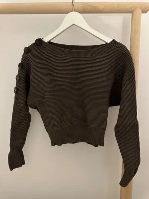 Brun ribbad stickad tröja Zara S - Snygg brun stickad tröja från Zara i storlek S. Tröjan har ribbad struktur och knappar längs ena axeln och ärmen. Modellen är lite croppad med bredare halsringning och långa ärmar.