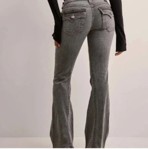 Grå True Religion Joey flare jeans - Säljer ett par grå  True Religion Joey jeans med låg midja och flare-ben. Snygga detaljer med stora fickor bak och silverfärgade knappar. Klassisk jeanslook med markerade sömmar och cool faded effekt. Perfekta för dig som gillar statement-jeans.