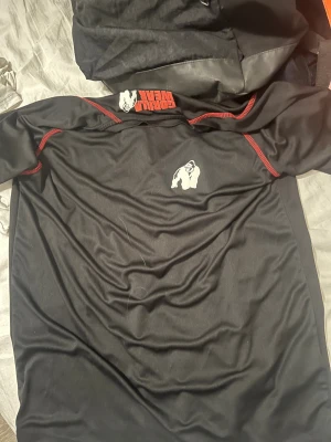 Svart Gorilla Wear t-shirt  - Säljer en svart t-shirt från Gorilla Wear med coolt gorilla-tryck på bröstet och röda kontrastsömmar. Materialet är mjukt och syntetiskt, perfekt för dig som gillar att träna och vill ha en sportig look. Passformen är normal och t-shirten har korta ärmar.