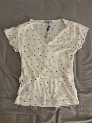 Vit blommig omlottblus från Fullamoda. - Supersöt vit omlottblus från Fulana med små röda blommor och diskreta vertikala ränder. Blusen har korta, lite volangiga ärmar och v-ringad hals. Perfekt för dig som gillar romantisk och feminin stil.