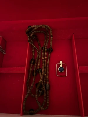 Brunt träpärlhalsband & silverberlock - Unikt halsband med flera rader små bruna träpärlor och mörka glaspärlor. Till halsbandet hör en modern silverfärgad berlock i rektangulär form med en rund blå sten i mitten. Perfekt för dig som gillar boho och retrovibbar.