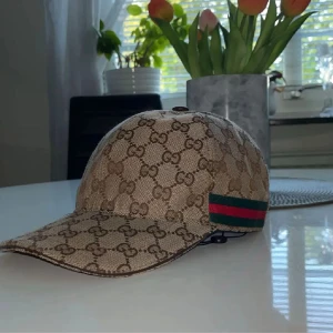 Gucci beige keps med GG-mönster - Snygg beige keps från Gucci med klassiskt GG-monogram över hela kepsen och en grön-röd rand på sidan. Justerbar rem baktill i brunt skinn. Materialet är bomull och polyester, insidan är mörkbrun. Perfekt för dig som vill ha en lyxig streetstyle.