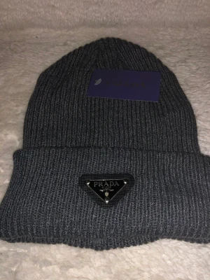 Grå ribbad mössa från Prada - Snygg grå mössa från Prada med ribbad stickning och uppvikt kant. På framsidan sitter en svart, triangelformad metallbricka med Prada-loggan. Mössan är tillverkad i mjukt stickat material och har en klassisk, rundad form som passar perfekt för kyliga dagar.