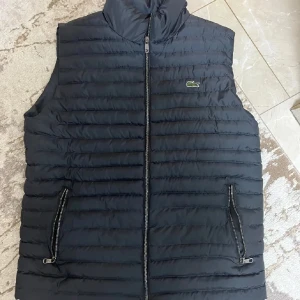 Svart dunväst från Lacoste - Snygg svart dunväst från Lacoste med quiltad design och dragkedja framtill. Västen har två fickor med dragkedja och klassisk Lacoste-logga på bröstet. Insidan är grön med tryckt Lacoste-mönster och stor logga. Den är knappast använd, eftersom de inte är min stil. Prisändringar endast vid snabb affär. Storlek M men passar även till S