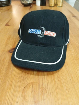 Svart keps Super Däck med brodyr - Svart keps med vit kant och broderad Super Däck-logga i blått, vitt och rött framtill. Klassisk form med böjd skärm och justerbar passform. Perfekt för dig som gillar bilar eller vill ha en cool accessoar.