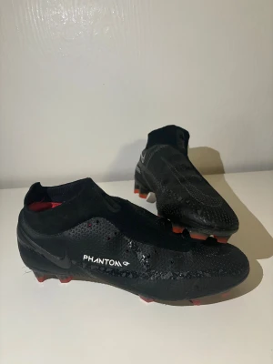 Nike Phantom GT svarta fotbollsskor - Nike Phantom GT fotbollsskor i svart med diskreta vita detaljer och röda dobbar. Skorna har en strumpliknande passform utan snörning, med syntetiskt material och texturerad yta för bättre bollkontroll. Snygg och modern design som sticker ut på planen. Pris kan diskuteras 