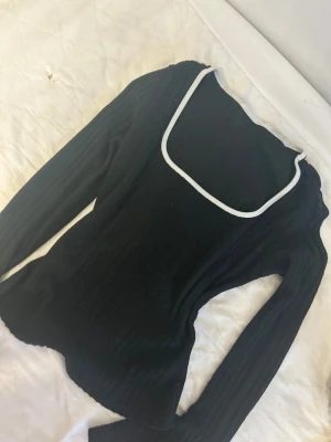 Svart ribbad långärmad topp med vit kant - Svart ribbad topp med lång ärm och vit kant runt den fyrkantiga halsringningen. Modellen är figurnära med utsvängda ärmslut och har en cool, modern vibe. Perfekt för dig som gillar stilrena detaljer och vill sticka ut med en enkel men snygg look.