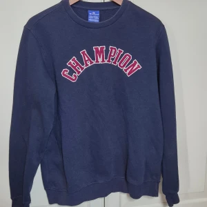 Mörkblå sweatshirt från Champion M - Mörkblå sweatshirt från Champion i storlek 2X-Large med klassisk röd och vit CHAMPION-text på bröstet. Tröjan har rund hals, långa ärmar och är tillverkad i mjuk bomullsmix. Perfekt för chill dagar och streetwear-stil. Sitter som en herr M.