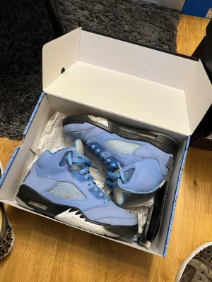 Nike Air Jordan 5 UNC blå sneakers - Nike Air Jordan 5 UNC sneakers i ljusblå mocka med svarta och vita detaljer på sulan. Skorna har snörning, meshpaneler på sidorna och en silverfärgad plös med Jumpman-logga. Insidan har mönstrat foder och texten 'HEART' och 'SOUL' på plösen. Perfekt för dig som vill sticka ut.