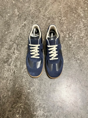 Maison Margiela Gats - Snygga sneakers i mörkblå mocka och skinn med vita snören och beige gummisula. Klassisk rund tå och platt sula, inspirerade av retro sportstil. Perfekt för dig som gillar stilrena och coola detaljer.