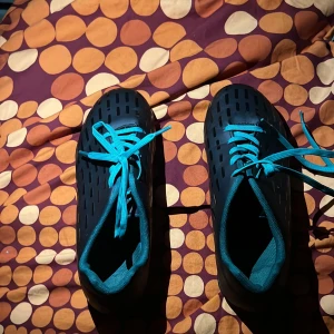 Svarta fotbollsskor med turkosa detaljer - Säljer ett par svarta fotbollsskor med turkosa snören och insida. Skorna har ventilerande hålmönster på ovansidan och en greppvänlig sula med turkosa inslag. Perfekt för dig som vill sticka ut på planen med en cool look.