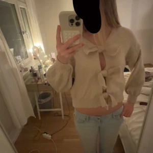 Beige kofta med rosetter från Shein - Superfin beige kofta från Shein i storlek S. Koftan är stickad och har tre stora rosetter framtill som stängning. Ballongärmar och ribbade muddar ger en trendig look. Perfekt att styla med jeans eller kjol för en söt vibe.