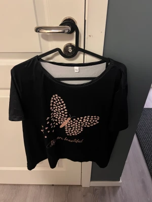 Svart topp med fjärilstryck XL - Svart topp i storlek XL med ett stort rosa fjärilstryck framtill och texten 'You are beautiful' under. Rund halsringning och korta ärmar. Mjuk och lätt bomullskänsla, perfekt för dig som gillar grafiska prints och chill stil.