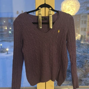 Lila stickad tröja från Polo Ralph Lauren - Snygg lila stickad tröja från Polo Ralph Lauren med klassisk v-ringning och kabelstickat mönster. Tröjan har långärmade ärmar och den ikoniska broderade loggan i gult på bröstet. Perfekt för dig som gillar stilrena och tidlösa plagg.