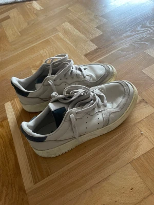 Adidas sneakers i vitt skinn - Säljer ett par Adidas sneakers i vitt skinn med mörkblå detaljer på hälen och tungan. Skorna har klassisk snörning, perforerade sidor och en platt, grov sula. Stilren och tidlös modell som passar perfekt till jeans eller shorts.