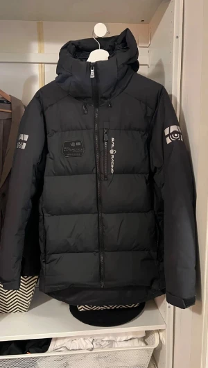 Saill racing vinterjacka - Svart dunjacka från Sail Racing med huva och coola loggor på ärmar och bröst. Jackan har dragkedja framtill, flera fickor och är tillverkad i vind- och vattenavvisande material med certifierat dun. Perfekt för kalla vinterdagar och har en clean, sportig look.