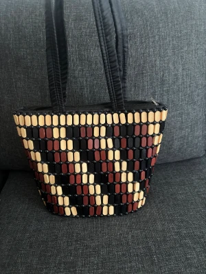 Handväska  - Unik handväska med geometriskt mönster i svart, beige och vinrött. Väskan har pärldetaljer och rejäla svarta handtag. Stängs med dragkedja upptill . 