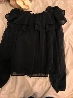 Svart blus med volang från Gina Tricot - Säljer en svart blus från Gina Tricot i storlek 158/164. Blusen har transparent tyg, volangdetaljer och långa ärmar med lätt puff. Perfekt för dig som gillar en romantisk och trendig stil. Materialet känns lätt och luftigt.