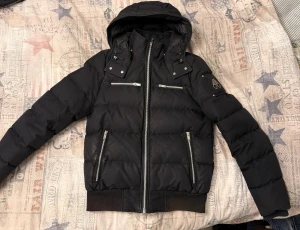 Svart pufferjacka från Moose Knuckles  - Svart pufferjacka från Moose Knuckles med huva, flera dragkedjefickor och ribbade muddar. Jackan har quiltad design och är tillverkad i polyester med dunfyllning. Snygga silverfärgade dragkedjor och logga på ärmen ger en cool vibe.