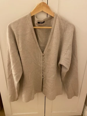 Beige kofta från Gina Tricot - Säljer en stilren beige kofta från Gina Tricot med v-ringad hals och dekorativa knappar framtill. Koftan har långärmad design och är gjord i mjukt stickat material som känns skönt mot huden. Perfekt att slänga på sig när det är lite kyligt ute.
