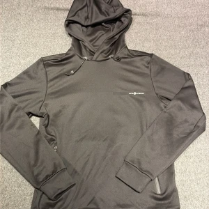 Svart hoodie från Sail Racing - Svart hoodie från Sail Racing med stilren design och diskret logga på bröstet. Tröjan har huva med snörning, långa ärmar och är tillverkad i mjuk polyester som känns skön mot huden. Perfekt för dig som gillar sportig och clean look.