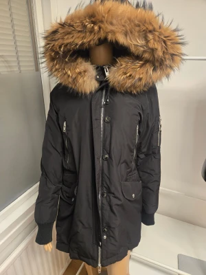Svart parkas från Rockandblue med pälskant - Svart parkas från Rockandblue med stor huva och lyxig pälskant i bruna toner. Jackan har dragkedja, knappar, snörning i midjan och flera fickor. Perfekt för kalla dagar med sin vadderade insida och långa modell, en inte så vanlig modell. Storlek 40  Använts sporadiskt under en vinter och har inget slitage eller så vad jag kan se när jag synar jackan noggrant. Pälsen är fin och fluffig. Matchande mössa från samma märke ingår (se sista bilden)
