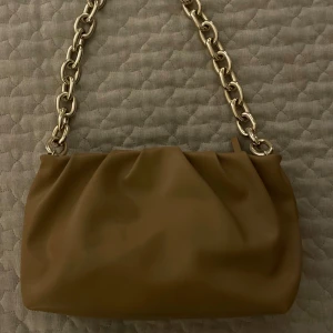 Beige axelväska med kedja från H&M - Snygg beige axelväska från H&M med mjukt, slätt material och en chunky silverfärgad kedja som axelrem. Väskan har ett rymligt fack med dragkedja och en stilren, modern look som passar till många outfits.