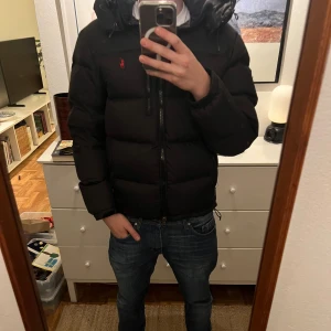 Polo Ralph Lauren jacka  - Svart dunjacka från Polo Ralph Lauren med huva och röd logga på bröstet. Jackan har quiltad design, dragkedja framtill och praktiska fickor med dragkedja. Perfekt för kalla dagar och har en clean, modern look som funkar till jeans och sneakers.