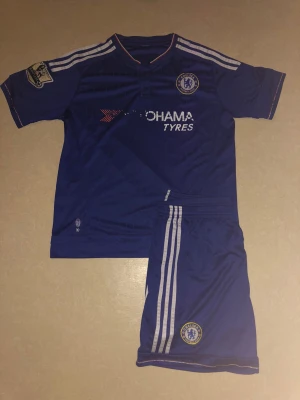 Chelsea FC blå fotbollströja och shorts - Chelsea FC matchtröja och shorts i blått med vita ränder på axlar och sidor. Tröjan har klubbmärke på bröstet, Premier League-märke på ärmen och sponsortryck framtill. Tillverkad i lätt polyester, perfekt för träning eller match.