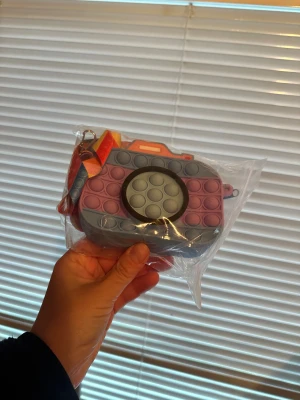 Pop it kameraformad axelväska Pop It Väska - Kamera - Säljer en unik axelväska i form av en kamera med pop it-detaljer. Väskan har en cool mix av grått, lila och orange samt ett färgglatt axelband i rött, orange och gult. Materialet är mjuk syntet och väskan stängs med dragkedja. Perfekt för dig som gillar lekfulla accessoarer.Rolig Pop It väska i form av en kamera. Perfekt för barn. Både för pojkar och flickor 