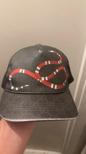 Gucci svart keps med röd orm - Snygg svart keps från Gucci med mesh på sidorna och bak, samt justerbar rem. Framtill pryds kepsen av en röd, vit och svart orm som slingrar sig över den klassiska GG-monogrammönstrade ytan. Perfekt för dig som vill sticka ut med en lyxig streetstyle-accessoar.