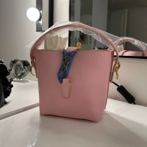 Rosa  handväska i skinn - Säljer en superfin ljusrosa handväska med klassisk logga framtill i metall. Väskan har guldfärgade detaljer, kort handtag och är tillverkad i slätt skinn. Perfekt storlek för det viktigaste och ger en söt touch till din outfit.