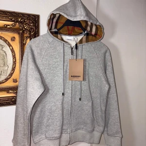 Grå Burberry hoodie med dragkedja - Snygg grå hoodie från Burberry med klassiskt rutigt foder i huvan. Tröjan har dragkedja framtill, snörning vid huvan och två fickor. Perfekt för dig som vill ha en stilren och lyxig streetwear-look.
