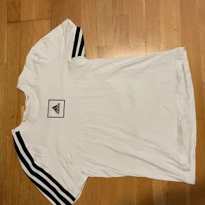 Vit Adidas t-shirt med svarta ränder - Vit t-shirt från Adidas med klassiska svarta ränder på axlarna och ett Adidas-logotyptryck på bröstet. Rund hals och korta ärmar. Skön och mjuk bomull som passar perfekt till en sportig stil.
