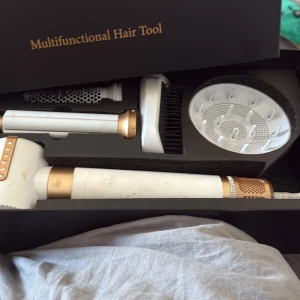 Multifunctional Hair Tool - Multifunktionellt hårverktyg med flera utbytbara tillbehör för styling, borstning och torkning. Elegant design i vitt och guld. Viss smuts och slitage syns på handtaget, men produkten verkar komplett och funktionell.