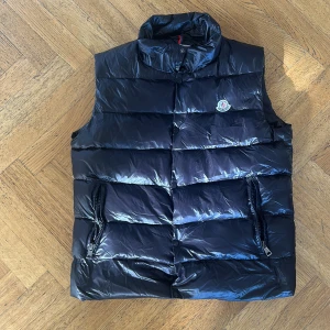 Svart dunväst från Moncler - Snygg svart dunväst från Moncler med glansig finish och klassisk logga på bröstet. Västen har hög krage, två dragkedjefickor och breda quiltade paneler. Perfekt för lager-på-lager och ger en clean streetstyle-vibe.