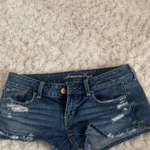 American Eagle jeansshorts låg midja - Snygga blå jeansshorts från American Eagle med låg midja och slitna detaljer framtill och baktill. Klassisk femficksmodell med gylf och knapp, samt coola slitningar för en trendig look. Perfekta till sommaren och lätta att matcha med allt.