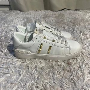 Valentino Rockstud vita sneakers - Snygga vita Valentino Rockstud sneakers i skinn med ikoniska guldiga nitar på sidorna och runt sulan. Klassisk rund tå, platt sula och snörning framtill. Perfekta för dig som vill ha en clean look med edgy detaljer.